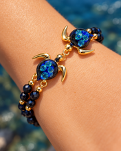 Hana Gold double sea turtle bracelet with dark navy glass and aqua inclusions worn on a wrist beside the ocean.
