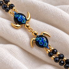 Close-up of Hana Gold double turtle bracelet with midnight-blue glass, large aqua inclusions and twin black bead strands on ivory linen.