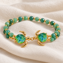Kalele Gold bracelet with two green glass turtles facing each other on ivory linen, with two rows of green beads.