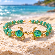Kalele Gold bracelet on a pale beach rock, with two facing green glass turtles and twin green bead strands.