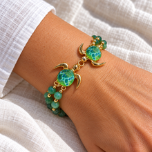 Kalele Gold double turtle bracelet with green glass shells and twin green bead strands worn on a wrist beside white linen.