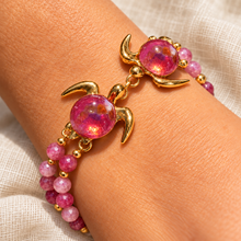 Kuipo Gold double turtle bracelet with pink glass shells and two rows of pink beads worn on a wrist against cream linen.