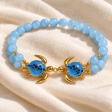 Mahalo Gold double turtle bracelet with sky-blue glass shells and two rows of pale-blue beads resting on cream linen.