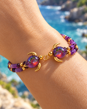 Malama Gold double sea turtle bracelet with plum-purple glass and purple bead strands worn on a wrist beside the ocean.