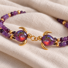 Close-up of two facing Malama Gold turtles with plum-purple glass, olive-green inclusions and warm red highlights on cream linen.
